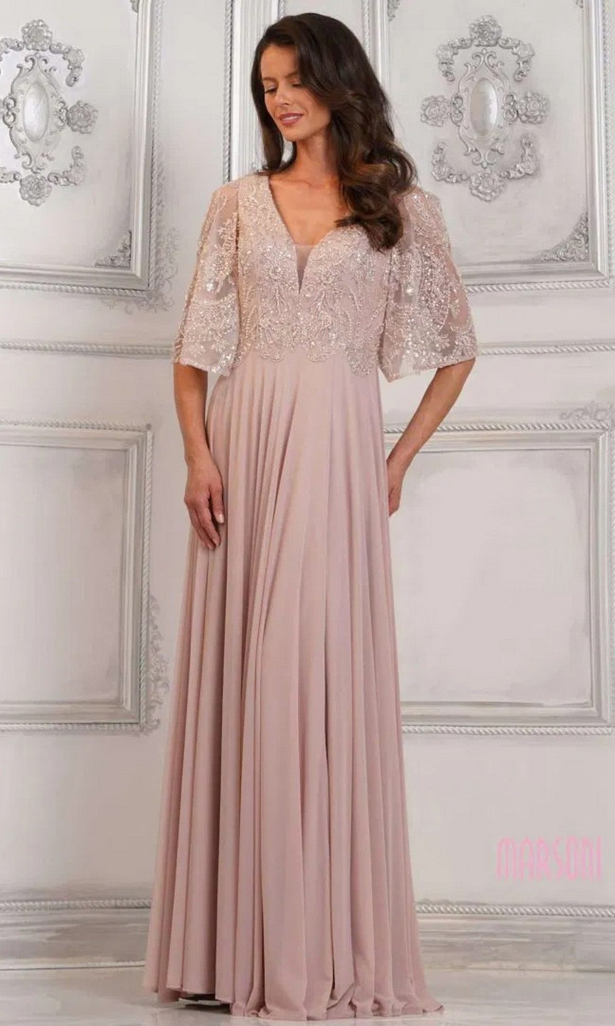 A woman in a mauve Marsoni MV1302 long formal dress with lace bodice and short sleeves stands before an ornate white wall.